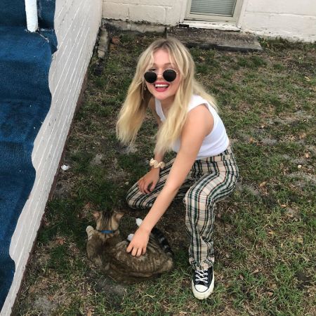 Emily Alyn Lind in a white top and pant petting a cat in her house.